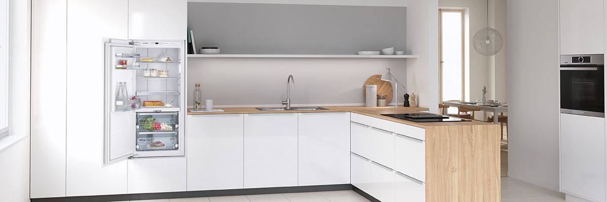Das Bild zeigt eine moderne, minimalistische Küche mit weißen Unterschränken und einer Holz-Arbeitsplatte. Im Hintergrund ist ein offener Regalschrank mit Geschirr und Küchenutensilien. Die Küche wirkt hell, geräumig und funktional, mit großem Fenster für natürliche Beleuchtung.